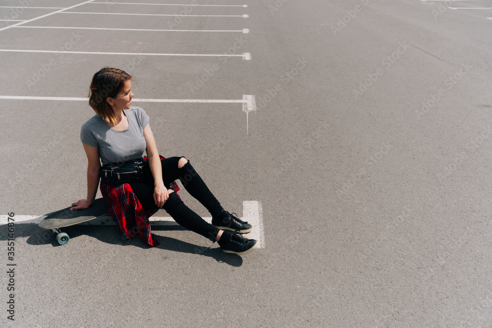 young woman sits with a longboard on the pavement in the parking lot in sunny weather. Active holiday on a summer day. Hobby sports skateboarding young woman sits with a longboard on the pavement in the parking lot in sunny weather. Active holiday on a summer day. Hobby sports skateboarding