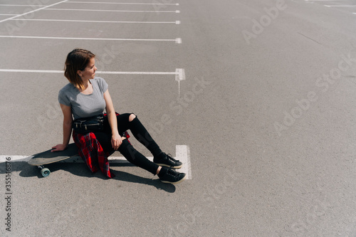 young woman sits with a longboard on the pavement in the parking lot in sunny weather. Active holiday on a summer day. Hobby sports skateboarding