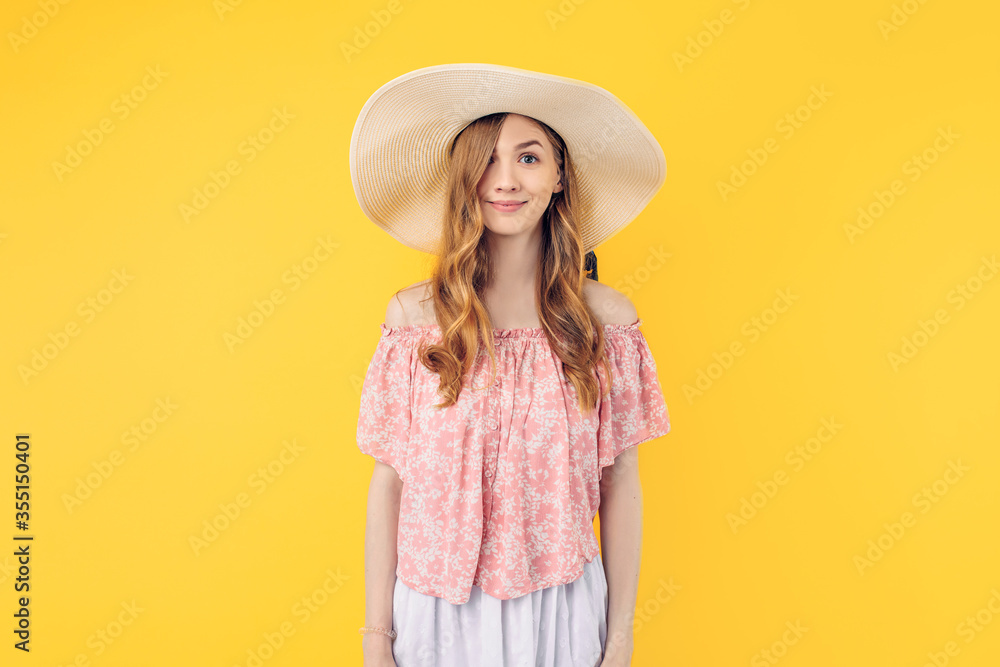 A happy attractive young woman in a summer dress and hat poses standing on an yellow background A happy attractive young woman in a summer dress and hat poses standing on an yellow background