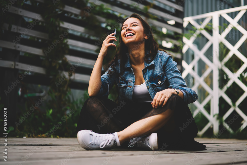 Happy young woman with gorgeous long hair laughing during mobile conversation via smartphone device sitting in lotus position.Cheerful female with mobile phone in hand having fun in free time outdoors Happy young woman with gorgeous long hair laughing during mobile conversation via smartphone device sitting in lotus position.Cheerful female with mobile phone in hand having fun in free time outdoors