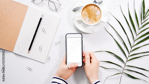 Flatlay on white work space desk with woman's hands holding white smartphone with white copyspace, coffee, planner and other office accessories. Mobile phone mockup.