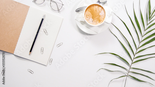 Flat lay white minimalistic office work space desk with copy space background. Work place with cappuccino cup coffee, notebook, glasses and palm branch.