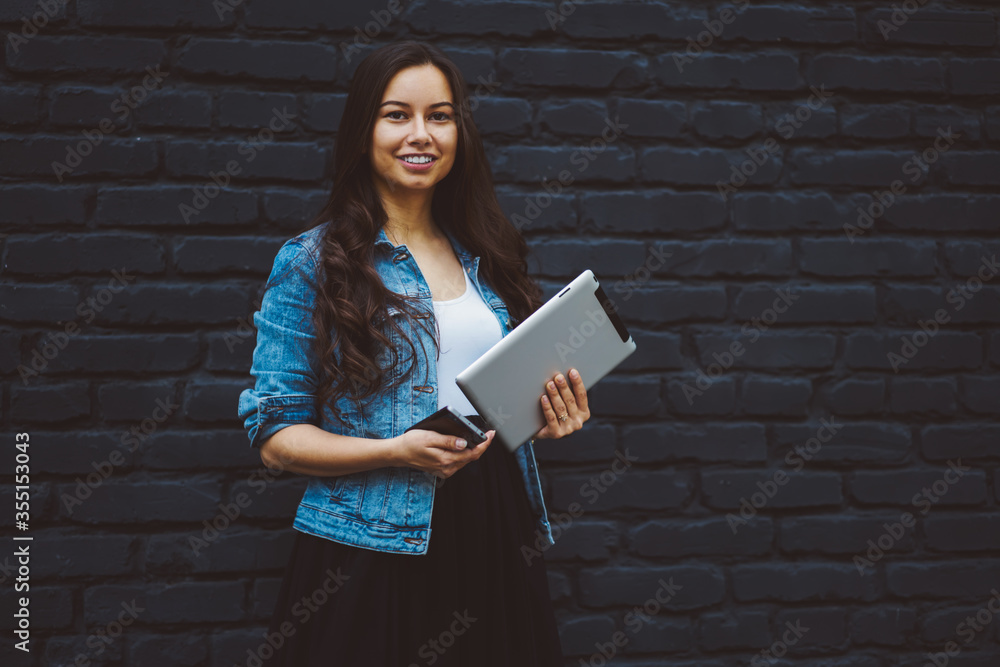 Half length portrait of beautiful smiling young woman blogger looking at camera while holding modern tablet and digital smartphone device in hands standing against promotional background Half length portrait of beautiful smiling young woman blogger looking at camera while holding modern tablet and digital smartphone device in hands standing against promotional background