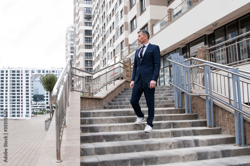 Handsome businessman with mobile in the city and looking away while walking outdoors with office building in the background Handsome businessman with mobile in the city and looking away while walking outdoors with office building in the background