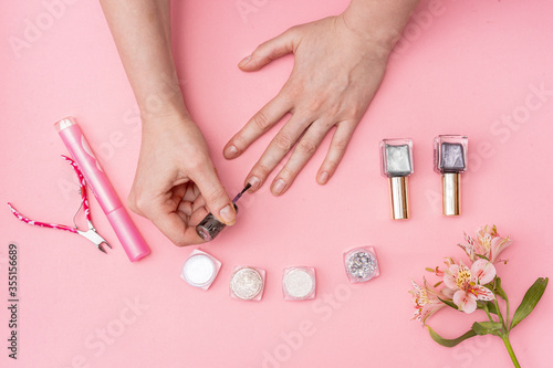 female hands paint nails on a pink background. next to it are cans of varnish and glitter boxes and scissors. in the corner is a pink flower