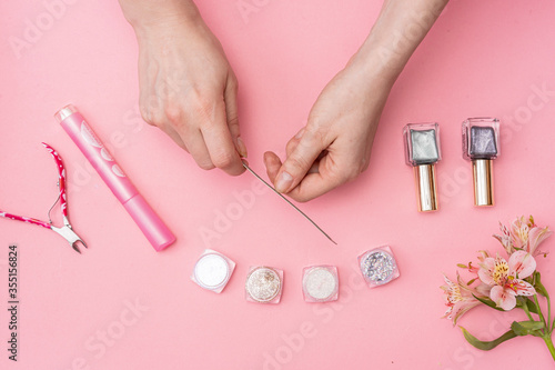 female hands use a nail file on a pink background. next to it are cans of nail polish and glitter boxes and scissors. in the corner is a pink flower
