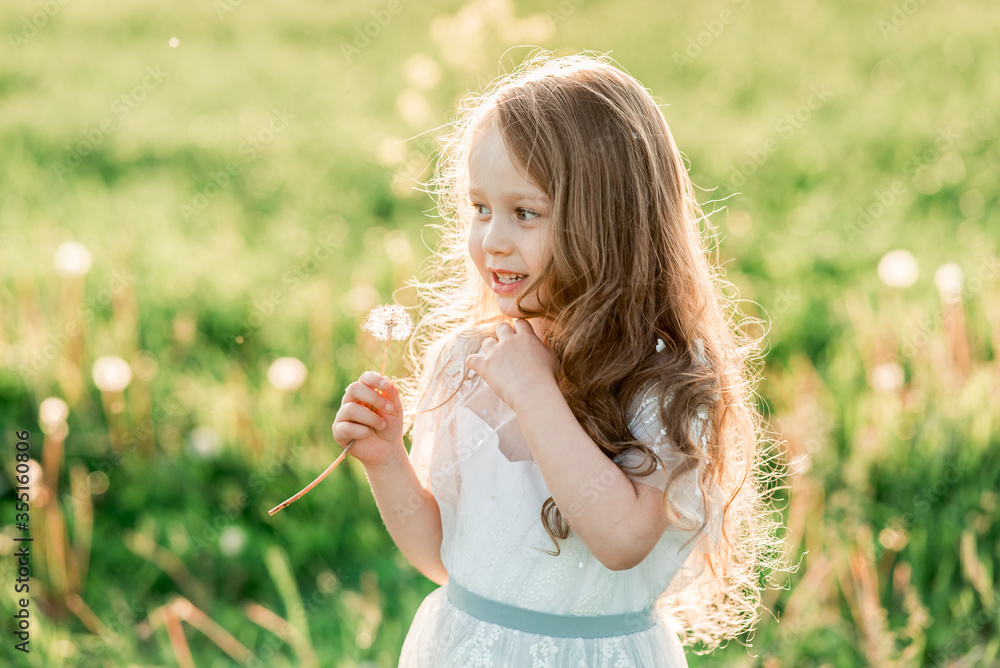 Portrait of cute curly little girl in white dress, staying in sunset meadow, smiling and holding dandelion. Healthy, enjoy countryside summer and happy childhood concept Portrait of cute curly little girl in white dress, staying in sunset meadow, smiling and holding dandelion. Healthy, enjoy countryside summer and happy childhood concept