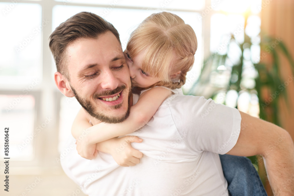 Fototapeta premium Attractive young father standing in the living room holds on his back a charming daughter at home. Smiling kid kisses dad on the cheek, playing a piggyback with dad. Father's day.