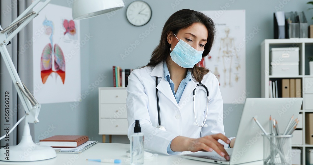 Caucasian young beautiful female doctor in white gown nd blue medical mask siting at desk in office and working at laptop computer Pretty woman medic typing on keyboard and describing illness symptoms Caucasian young beautiful female doctor in white gown nd blue medical mask siting at desk in office and working at laptop computer Pretty woman medic typing on keyboard and describing illness symptoms