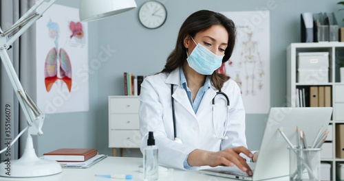 Caucasian young beautiful female doctor in white gown nd blue medical mask siting at desk in office and working at laptop computer Pretty woman medic typing on keyboard and describing illness symptoms
