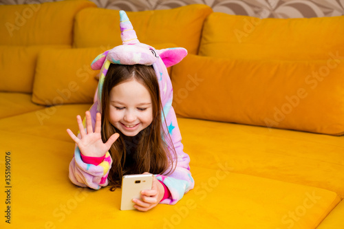 little girl in kigurumi smiles at home on the couch, taking a selfie, talking via video link.