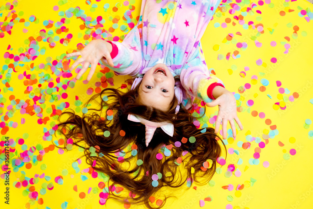 little girl with long hair wearing a kigurumi and headphones in the shape of a unicorn is happy catching confetti standing on a yellow background little girl with long hair wearing a kigurumi and headphones in the shape of a unicorn is happy catching confetti standing on a yellow background