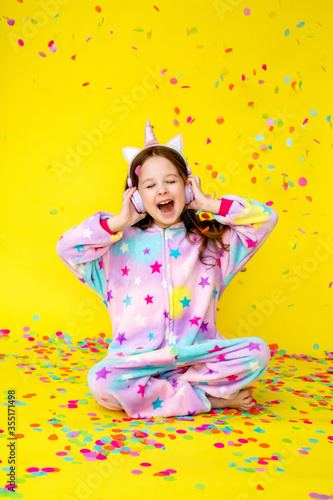 little girl with long hair wearing a kigurumi and headphones in the shape of a unicorn is happy catching confetti standing on a yellow background