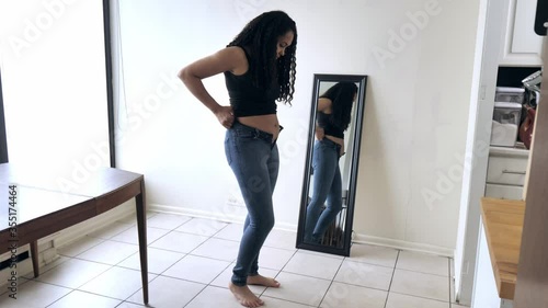Beautiful African American woman and mother stands in front of a mirror and attempts to try to close her tight fitting blue jeans pants after postpartum diet and weight loss exercise.
