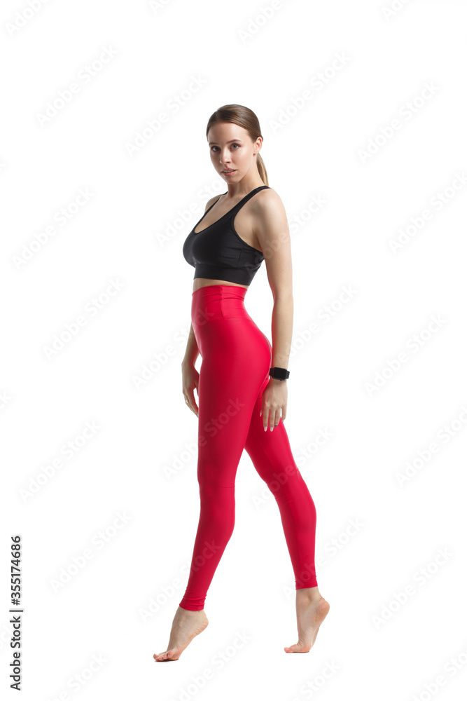 Slender athletic girl in sportswear a black top and pink leggings stands in full growth isolated on a white background.