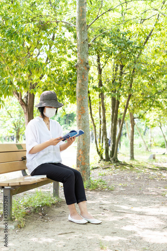 公園のベンチで帽子をかぶりマスクをして読書する1人の女性