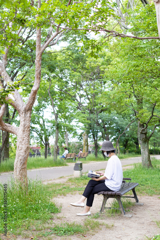 公園のベンチで帽子をかぶりマスクをして読書する1人の女性