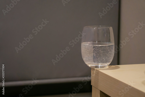 Fotografie A glass of water with bubbles inside is on the corner of light yellow wooden table in the grey room