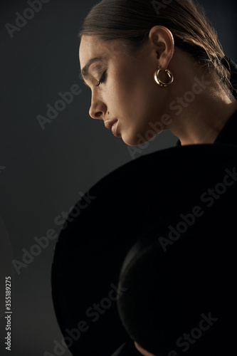 profile of a beautiful girl with closed eyes, she has a professional makeup, earrings in her ear, she and covered with a black hat on a dark background in the studio