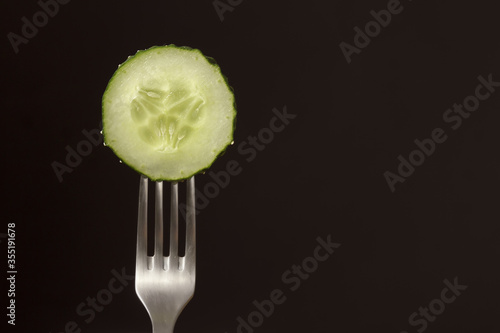 Slice of fresh green cucumber on fork on dark black background. silhouette of a fork.