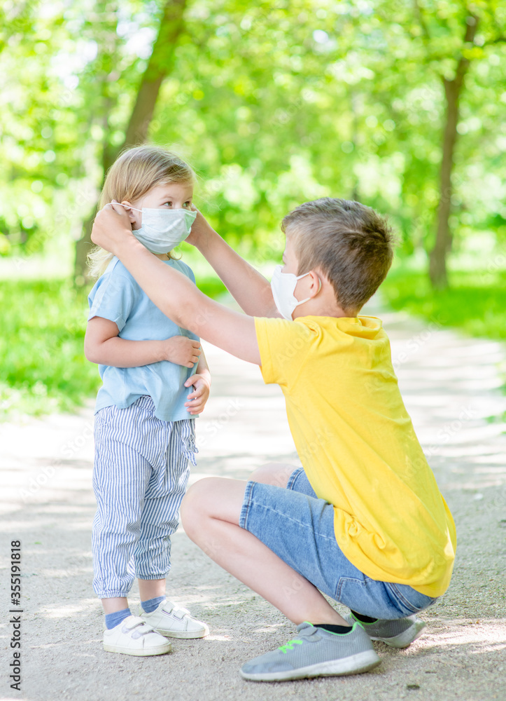 Brother takes care of his sister and helps her put on protective facemask while walking in a summer park Brother takes care of his sister and helps her put on protective facemask while walking in a summer park