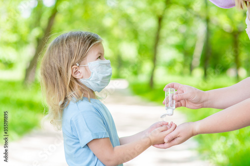 Mother applies sanitizer for cleaning baby hands in public place. Little girl wearing protective face mask looks at mom