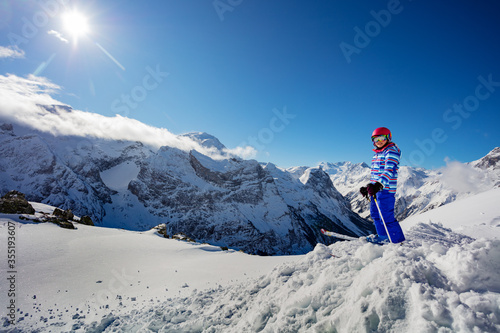 Cute ski young girl in colorful clothes stand over wide magnificent panorama of Alps tops on sunny winter day