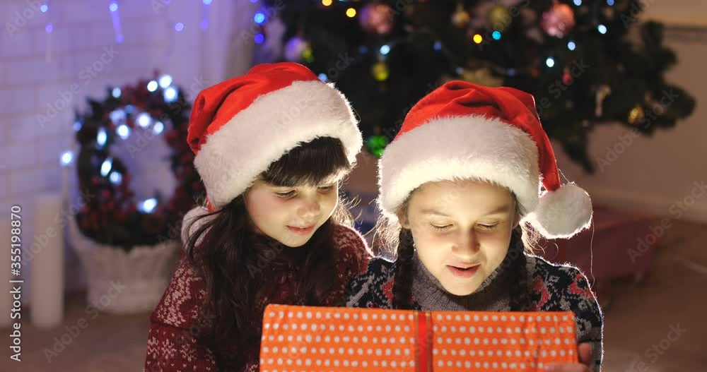 Two girls in warm sweaters and red Santa Claus hats open gifts under the Christmas tree. Portrait of sisters unpacking a festive orange box with a red ribbon from which light shines on their faces. Two girls in warm sweaters and red Santa Claus hats open gifts under the Christmas tree. Portrait of sisters unpacking a festive orange box with a red ribbon from which light shines on their faces.