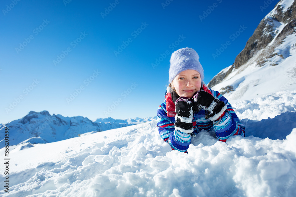 Happy smiling little girl lay in snow laying putting head on hands and smile wearing winter hat with mountain on background Happy smiling little girl lay in snow laying putting head on hands and smile wearing winter hat with mountain on background