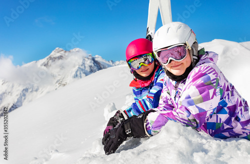 Close portrait of two girls lay in the snow over blue sky and high mountains with ski on background