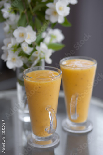 two yellow smoothie cocktails in high glasses on a glass breakfast table in the morning with white flowers. Side view. Summer Spring
