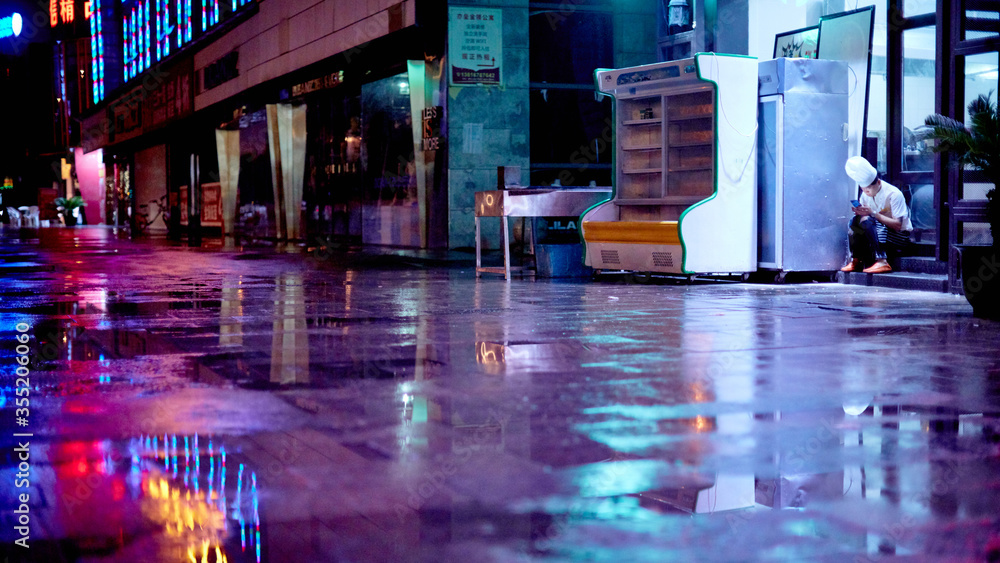 Chef takes a break out back of a Chinese restaurant in Shanghai, China. The cook sit on the step with his mobile phone on a rainy night, with coloured reflections on the ground from the lights