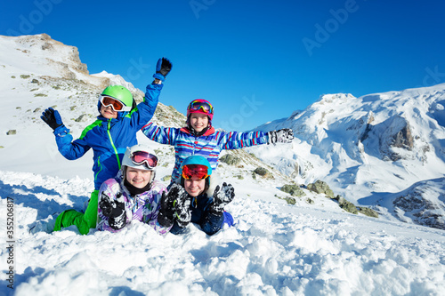 Group of four happy kids lay together in the snow on mountain top wear ski outfit smile and wave hands