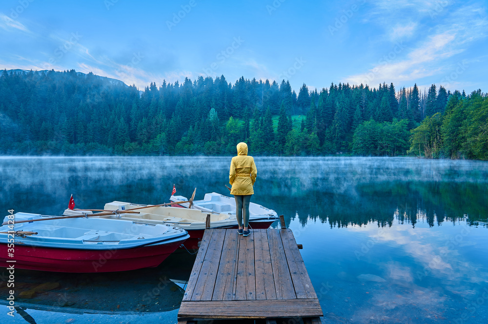 Fototapeta premium A girl in yellow coat is standing and posing on the wooden dock near boats on the foggy lake Karagol / Karagöl. Landscape taken in Savsat / Şavşat, Artvin, Black Sea / Karadeniz region of Turkey.