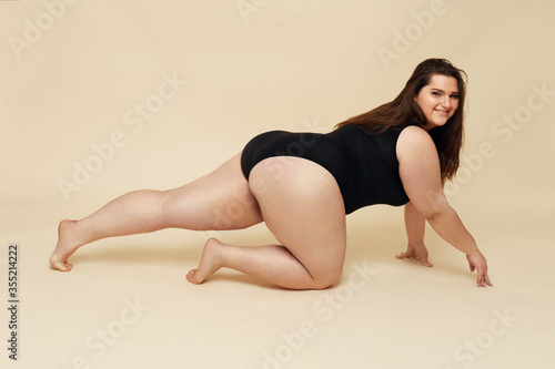 Plus Size Model. Crawling Big Woman In Black Bodysuit Portrait. Brunette Posing On Beige Background. Body Positive Concept.