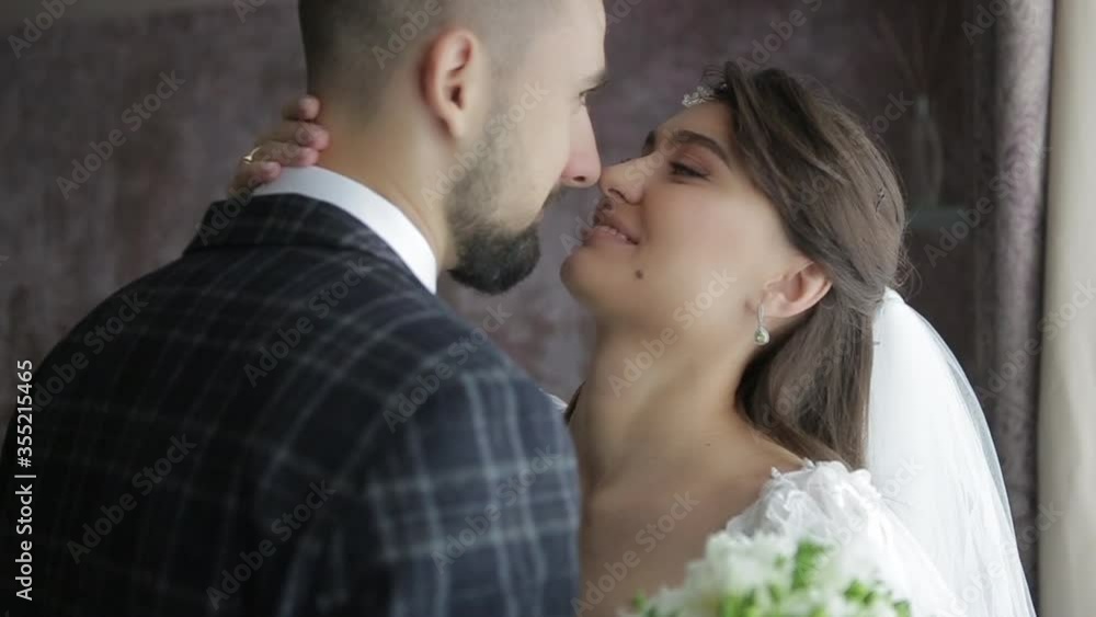Close-up shot of beautiful bride with diadem on her head, with bouquet of flowers and in a chic white wedding dress kissing her beloved groom in a plaid suit in the morning in an expensive hotel room Close-up shot of beautiful bride with diadem on her head, with bouquet of flowers and in a chic white wedding dress kissing her beloved groom in a plaid suit in the morning in an expensive hotel room
