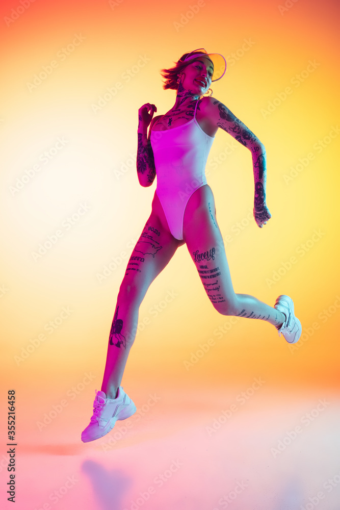 Young caucasian woman in swimsuit and beach hat jumping, running on gradient yellow background in neon. Beautiful model with tattoos. Human emotions, sales, ad concept. Resort and vacation, summertime Young caucasian woman in swimsuit and beach hat jumping, running on gradient yellow background in neon. Beautiful model with tattoos. Human emotions, sales, ad concept. Resort and vacation, summertime