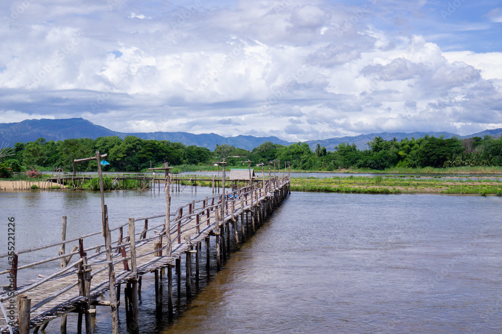 The wooden bridge over the river has a forested background. Mountains and blue skies