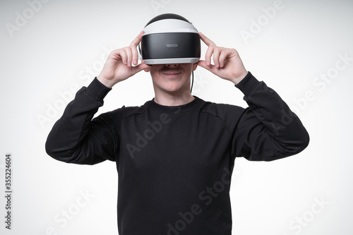 Young man in VR goggles watches a pleasant video and smiles widely, trying to adjust his glasses with hands, white background, studio