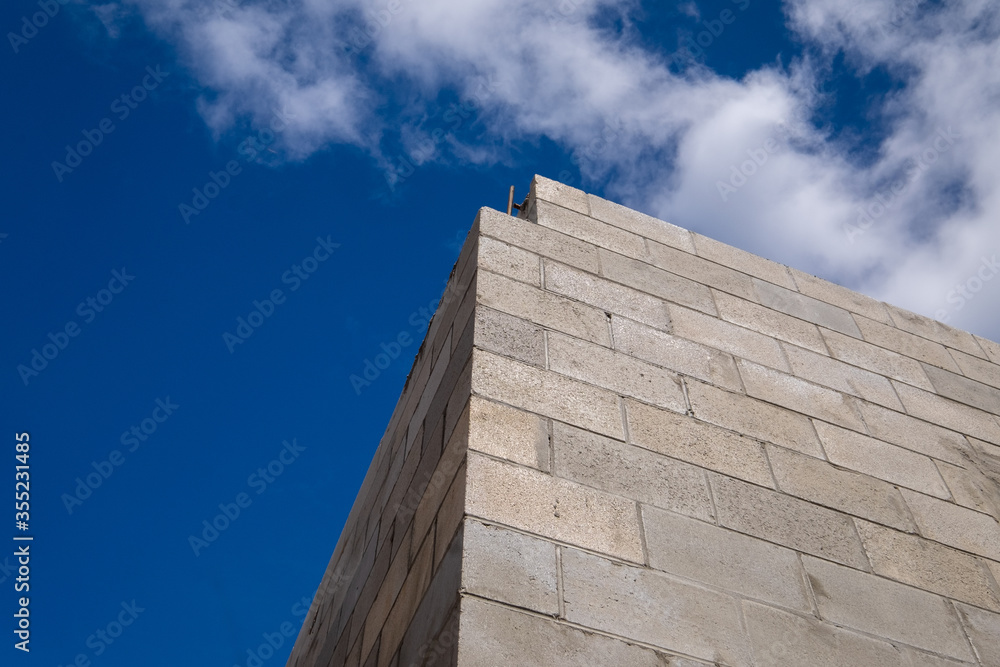 Corner view of a new large concrete brick building under construction. The stone is a tan colour with a brown mortar. The view is upward to the bright blue sky with some white fluffy clouds. Corner view of a new large concrete brick building under construction. The stone is a tan colour with a brown mortar. The view is upward to the bright blue sky with some white fluffy clouds.