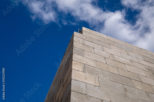 Corner view of a new large concrete brick building under construction. The stone is a tan colour with a brown mortar. The view is upward to the bright blue sky with some white fluffy clouds. 