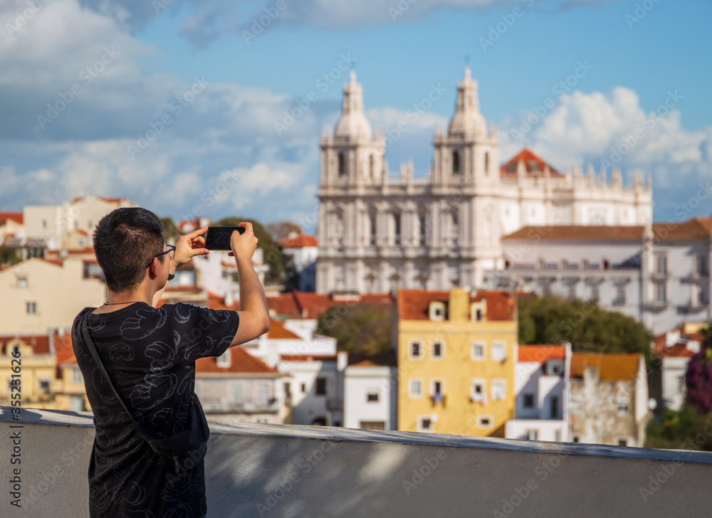Young Asian tourist in Lisbon Portugal - he is holding his cell phone and taking a picture of Church - Monastery of Sao Vicente de Fora and Alfama district.