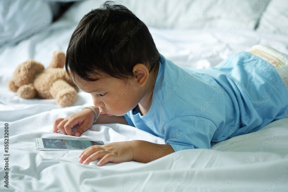 Asian boy about 2 years old, watching and playing mobile phone on the white bed. Asian boy about 2 years old, watching and playing mobile phone on the white bed.