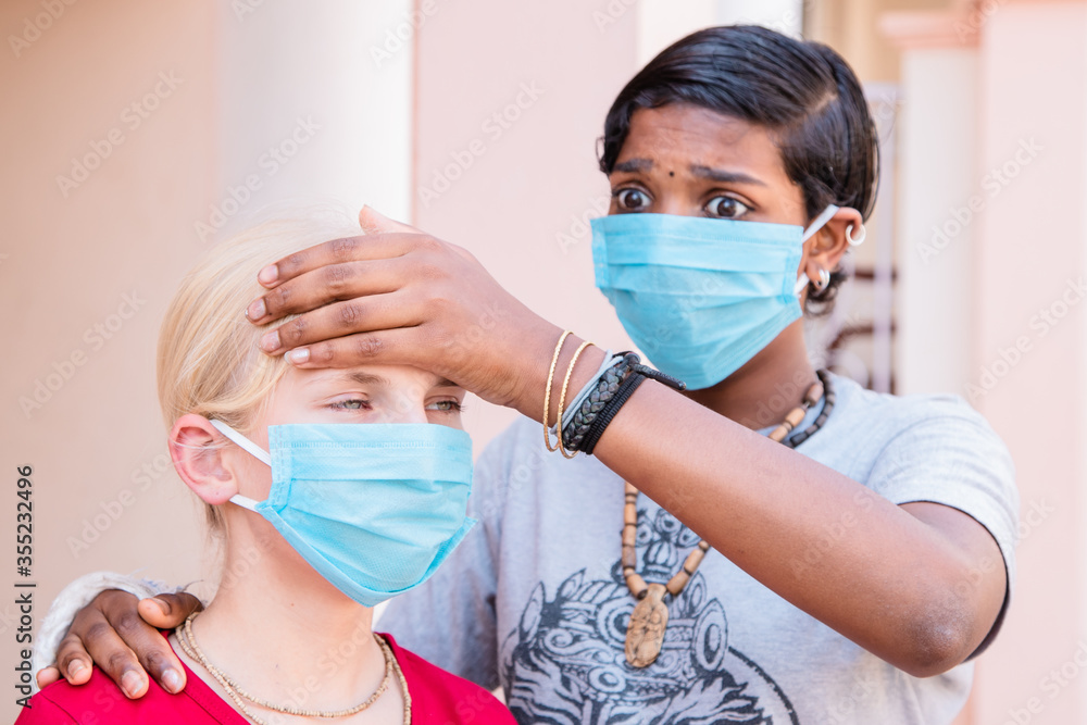 portrait of two Caucasian and African girls in protective medical masks, coronavirus concept. alarmed teenage girl checks her friend's temperature. multinational friendship