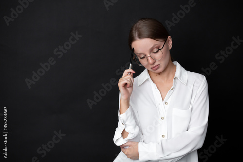 Beautiful woman businesswoman in glasses on a black background with a pen thinks looking down. Blank for advertising layout. Place for your text.