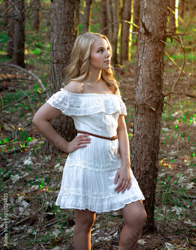 Blonde haired model with blue eyes in a white dress posing for a summer photo shoot in a forest. © Courtney