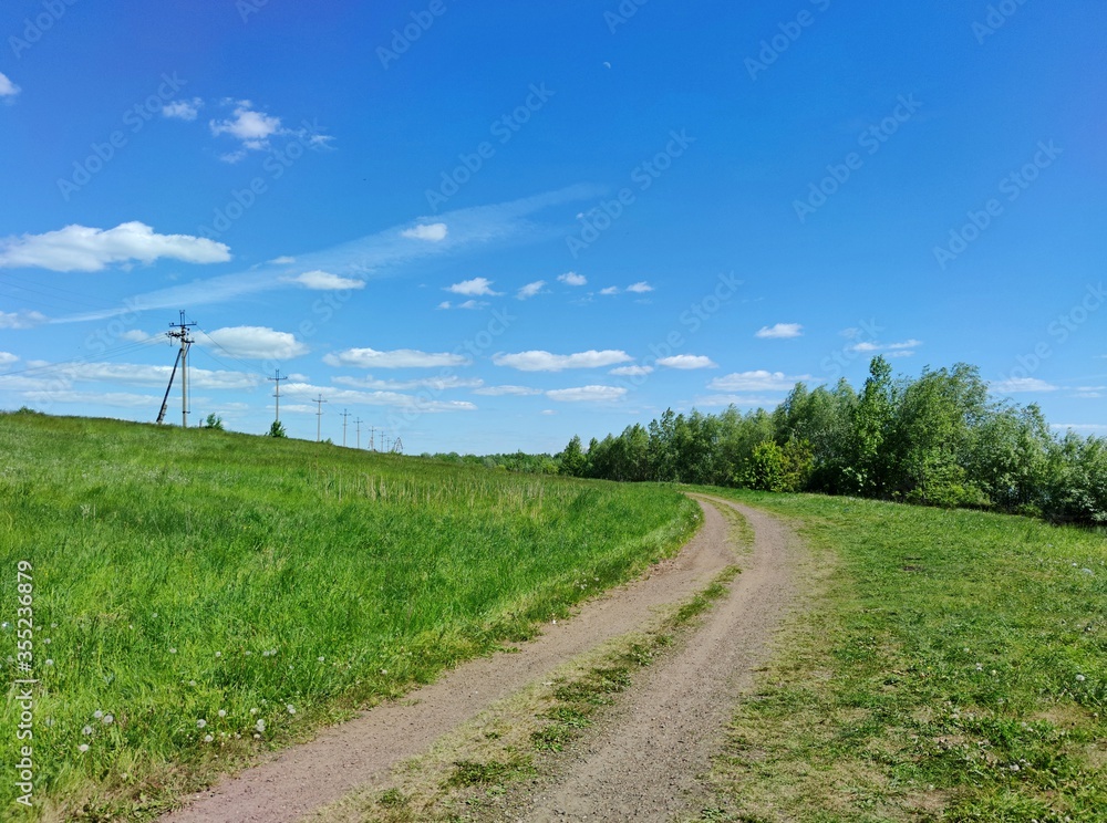 country road on a slope with green grass near a power line against a blue sky with clouds on a sunny day