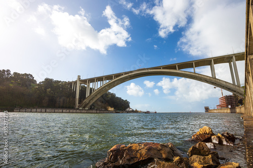 Porto Portugal: Ponte da Arrabida bridge on Douro river on a sunny day with brown rocks in the foreground and white clouds in a blue sky.