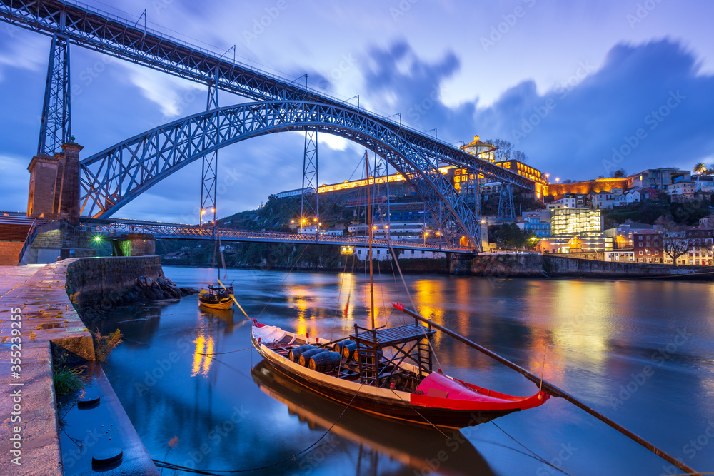 Naklejka premium Dawn in Porto, Portugal: The old iron bridge on Douro river, Luis I Ponte, at night, before sunrise. Traditional Portuguese boat used for transporting barrels of port wine in the foreground.