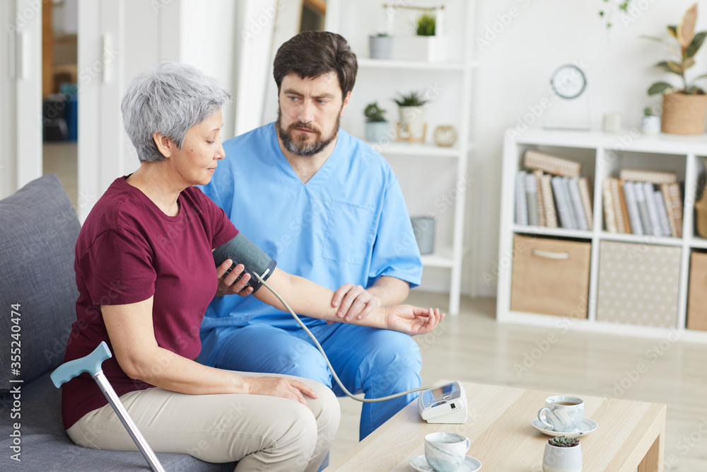 Obraz premium Male doctor measuring blood pressure of senior woman while they sitting on sofa at nursing house